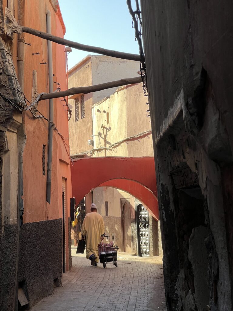 Skinny streets of Marrakesh, with a local man in a traditional robe and hat, pulling a cart behind him down the alley.